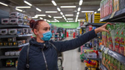 Woman wearing a blue mask reaches for packaged beverages on a supermarket shelf